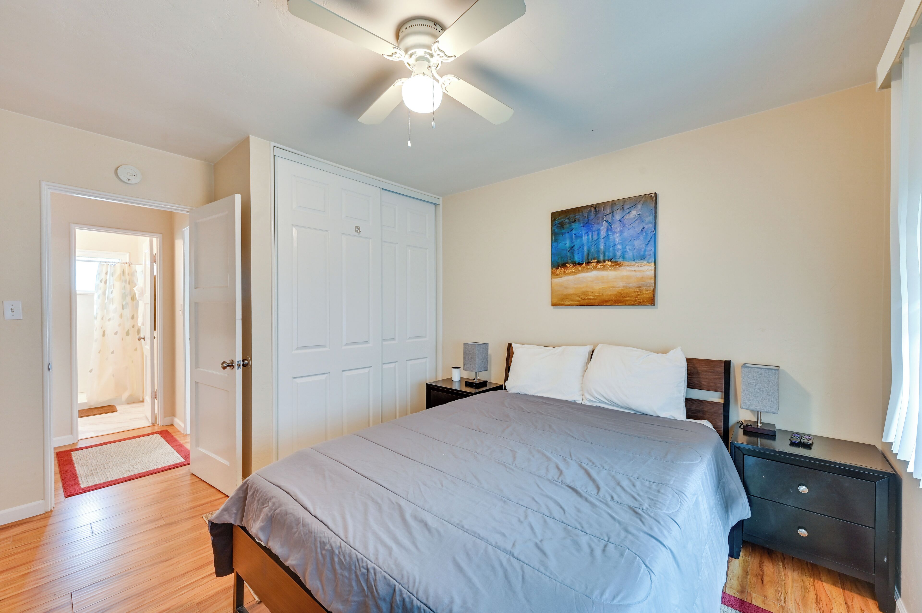 A bedroom with a bed, nightstands, a ceiling fan, a door, and a painting on the wall.