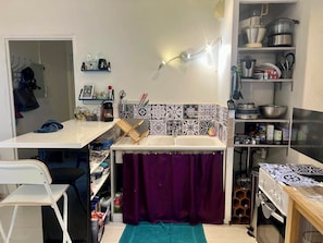 Pieni, käytännöllinen keittiö, jossa on valkoinen työtaso, pesuallas, violetti verho sekä erilaisia keittiövälineitä ja -laitteita.