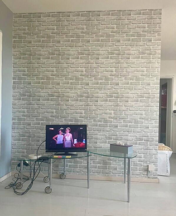 Olohuone, jossa on tiilikuviolla koristeltu seinä, lasinen sohvapöytä ja jalustalla oleva televisio.