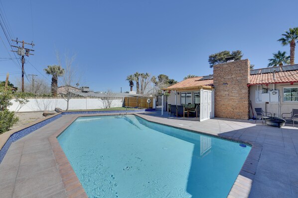 Une piscine avec un bord carrelé, un espace patio doté de chaises et un petit bâtiment surmonté d’un toit en tuiles.