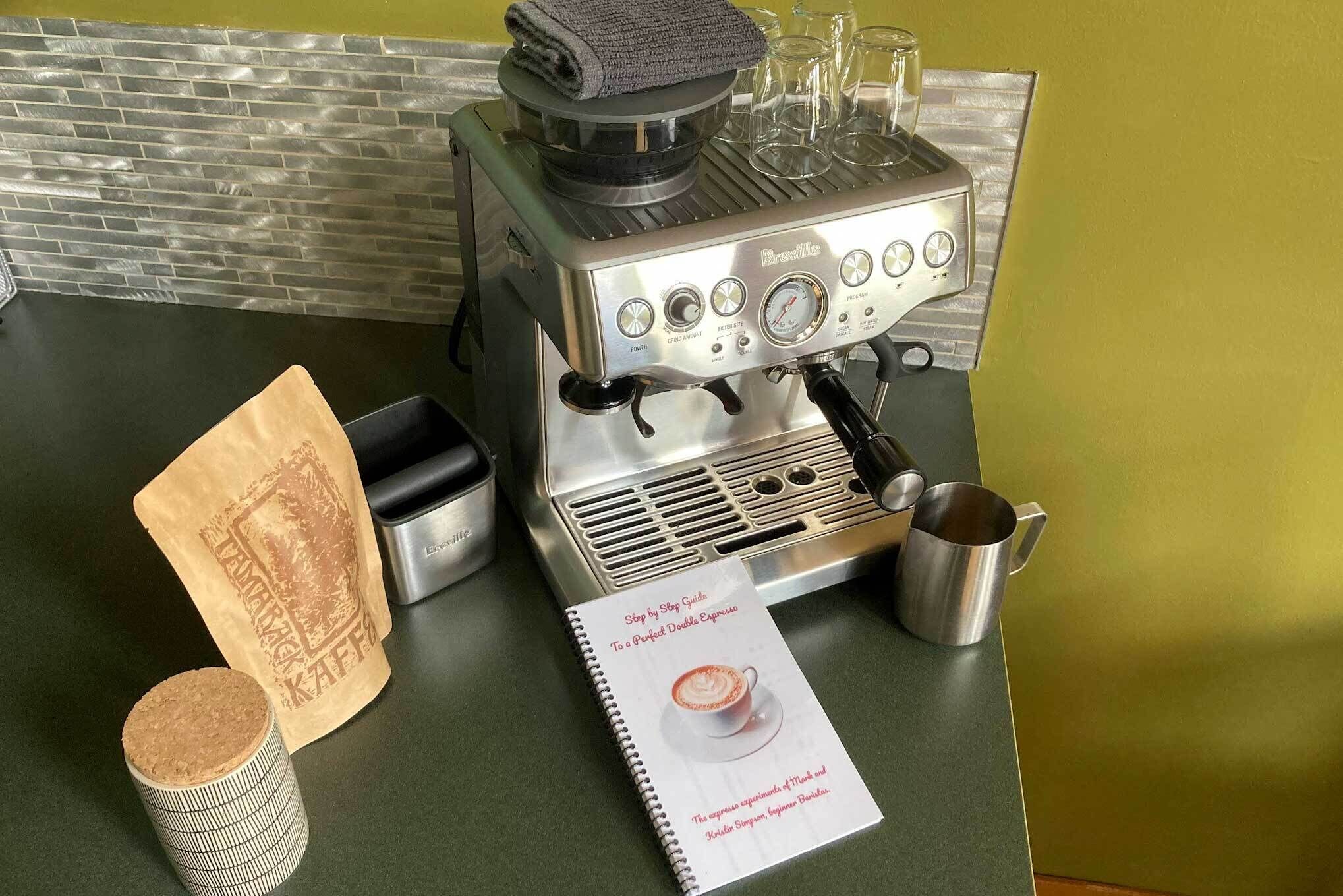 A Breville espresso machine on a kitchen counter with a coffee bag, a metal pitcher, and a guidebook.