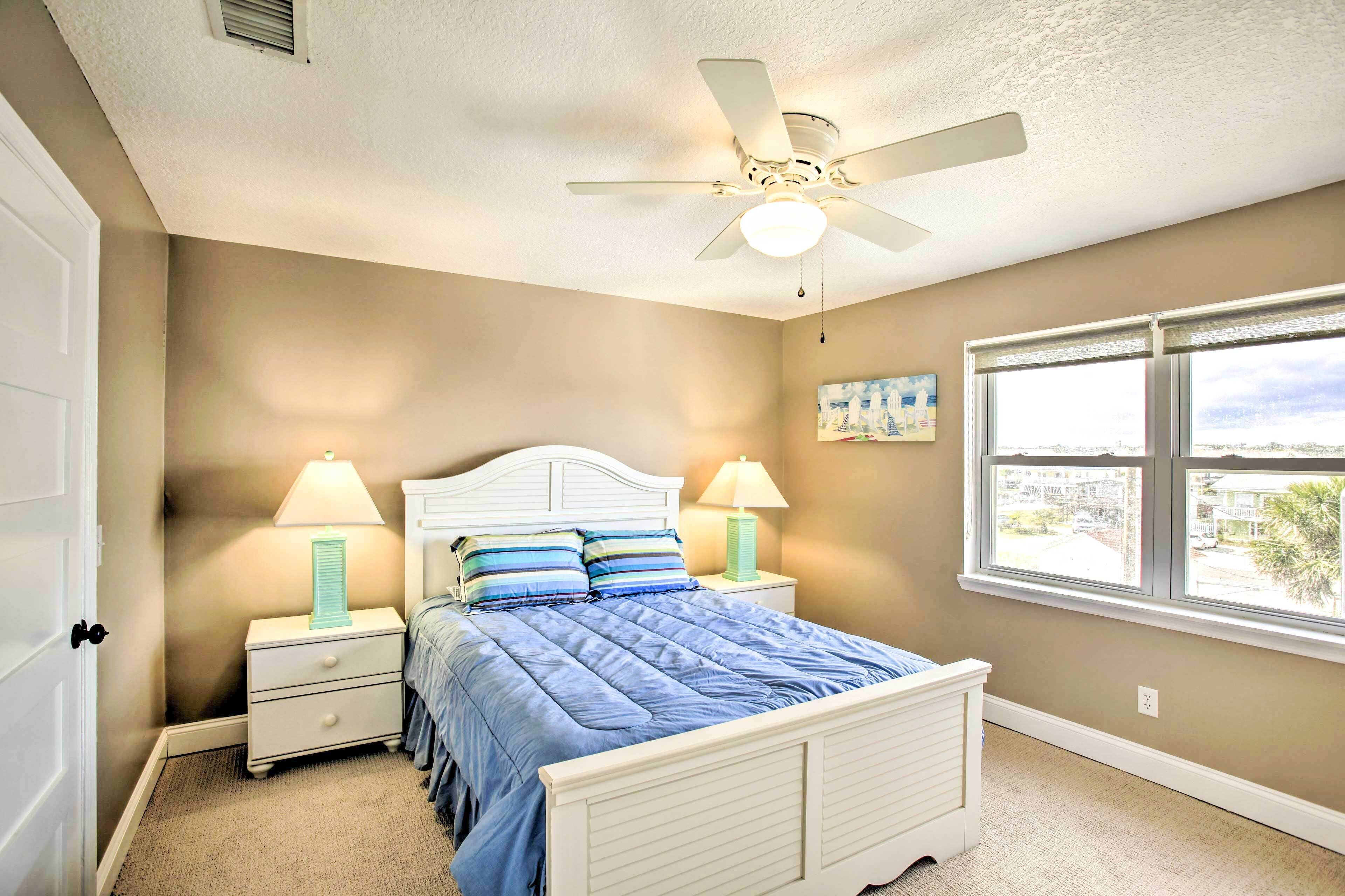 A bedroom with a bed, nightstands, lamps, a ceiling fan, and a window with a city view.