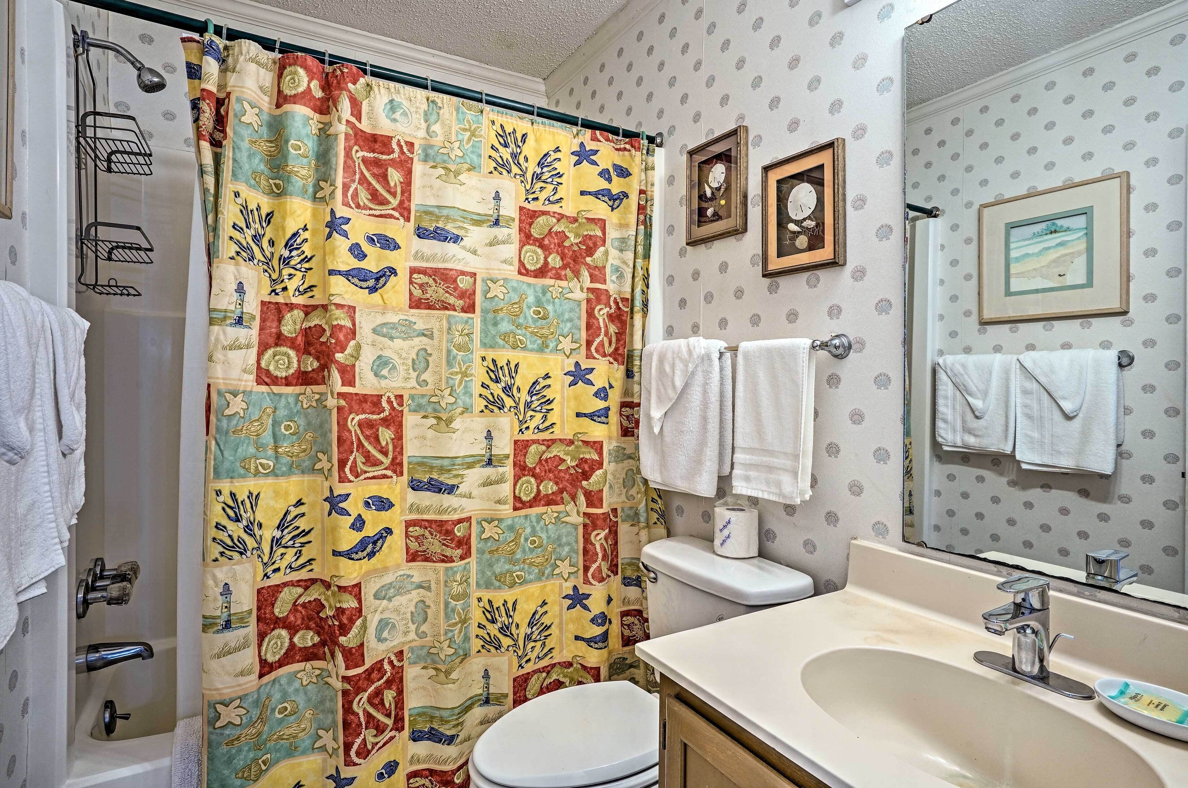 Une salle de bain avec un rideau de douche matelassé aux couleurs vives, des toilettes blanches, un lavabo avec un miroir et deux tableaux encadrés accrochés au mur.