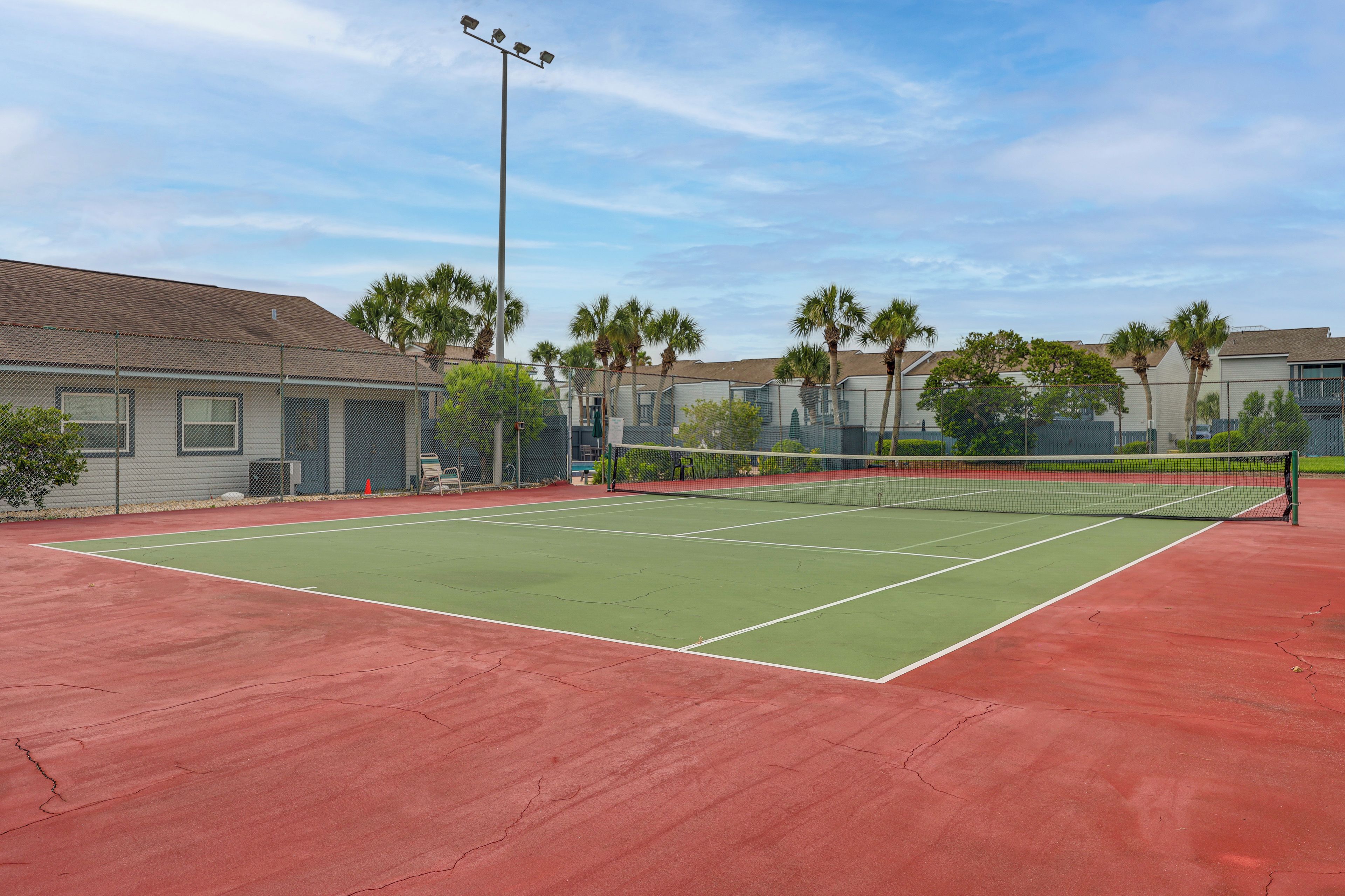 A tennis court with a net, surrounded by a fence, and palm trees in the background.