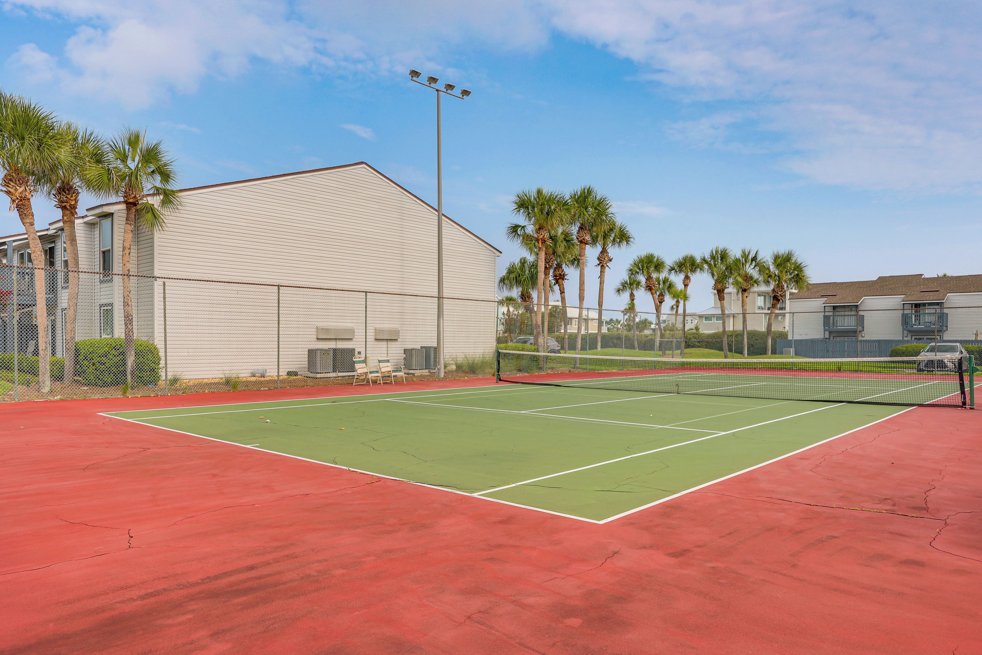 An outdoor tennis court with a net, surrounded by a fence, palm trees, and buildings in the background.