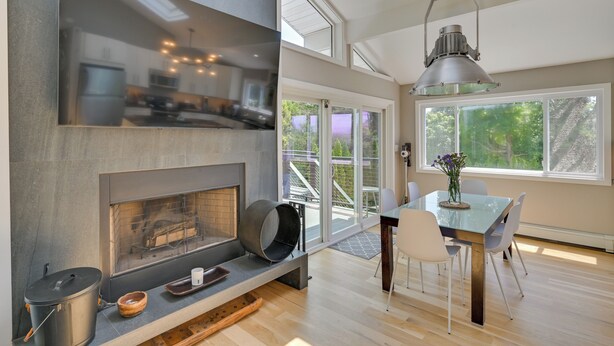 A modern kitchen with a fireplace, dining area with a table and chairs, and large windows offering a view of the outdoors.