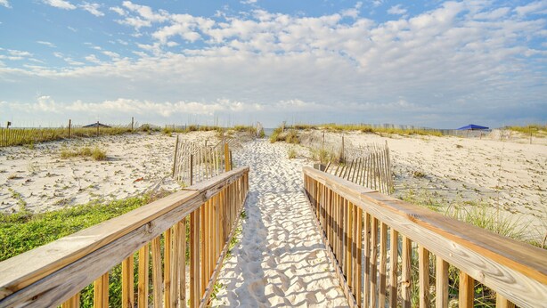 Ein hölzerner Steg führt zu einem Strand mit Sand und vereinzelter Vegetation.