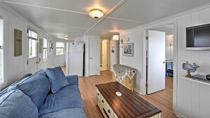 Une pièce avec un canapé bleu, une table basse en bois et une petite table recouverte d’une nappe en dentelle. On y trouve des portes menant à d’autres pièces et un téléviseur fixé au mur.