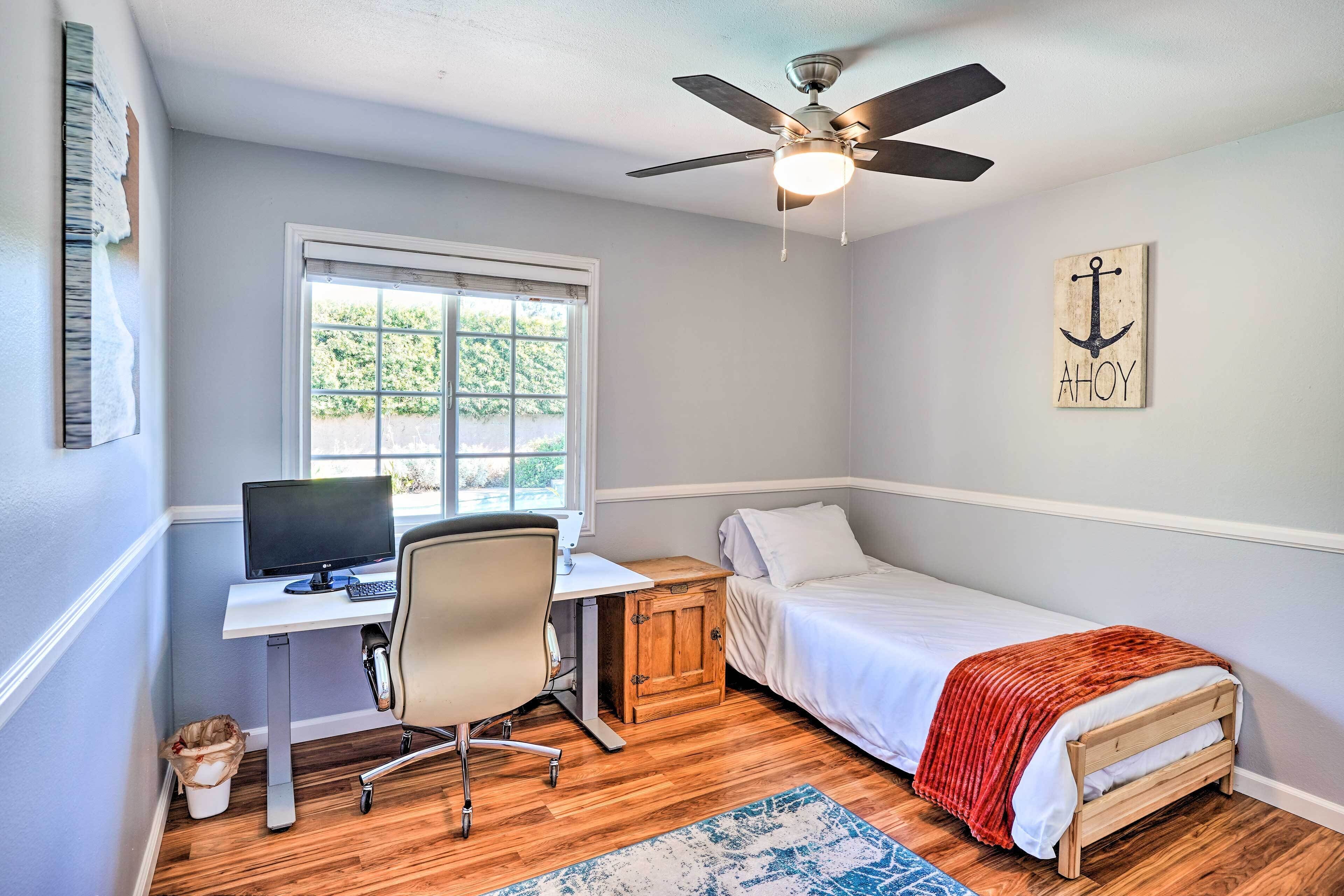 A bedroom with a single bed, a desk with a computer, a chair, a wooden cabinet, and a ceiling fan.