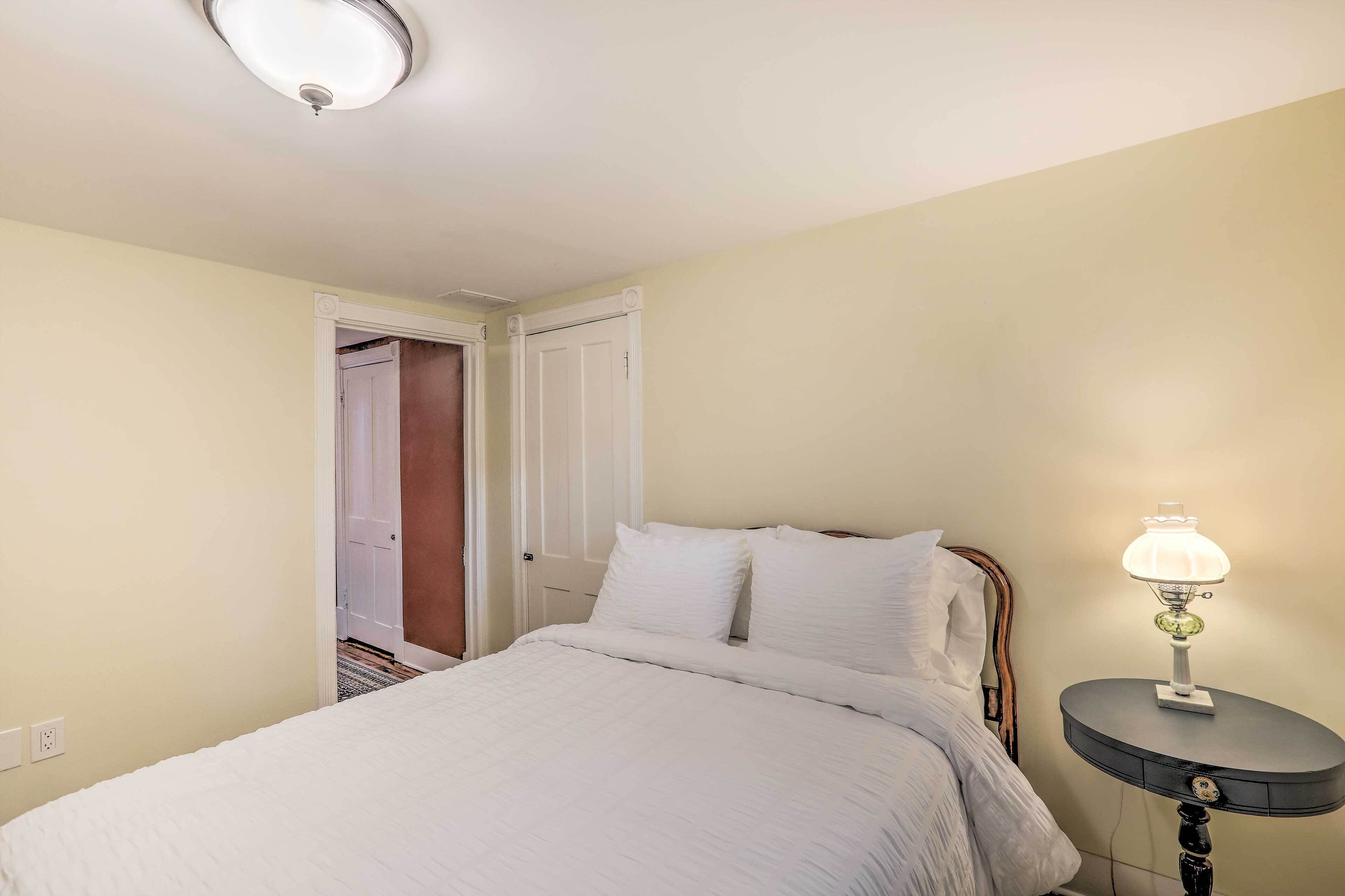 A bedroom with a bed, bedside table, lamp, and a door leading to another room.