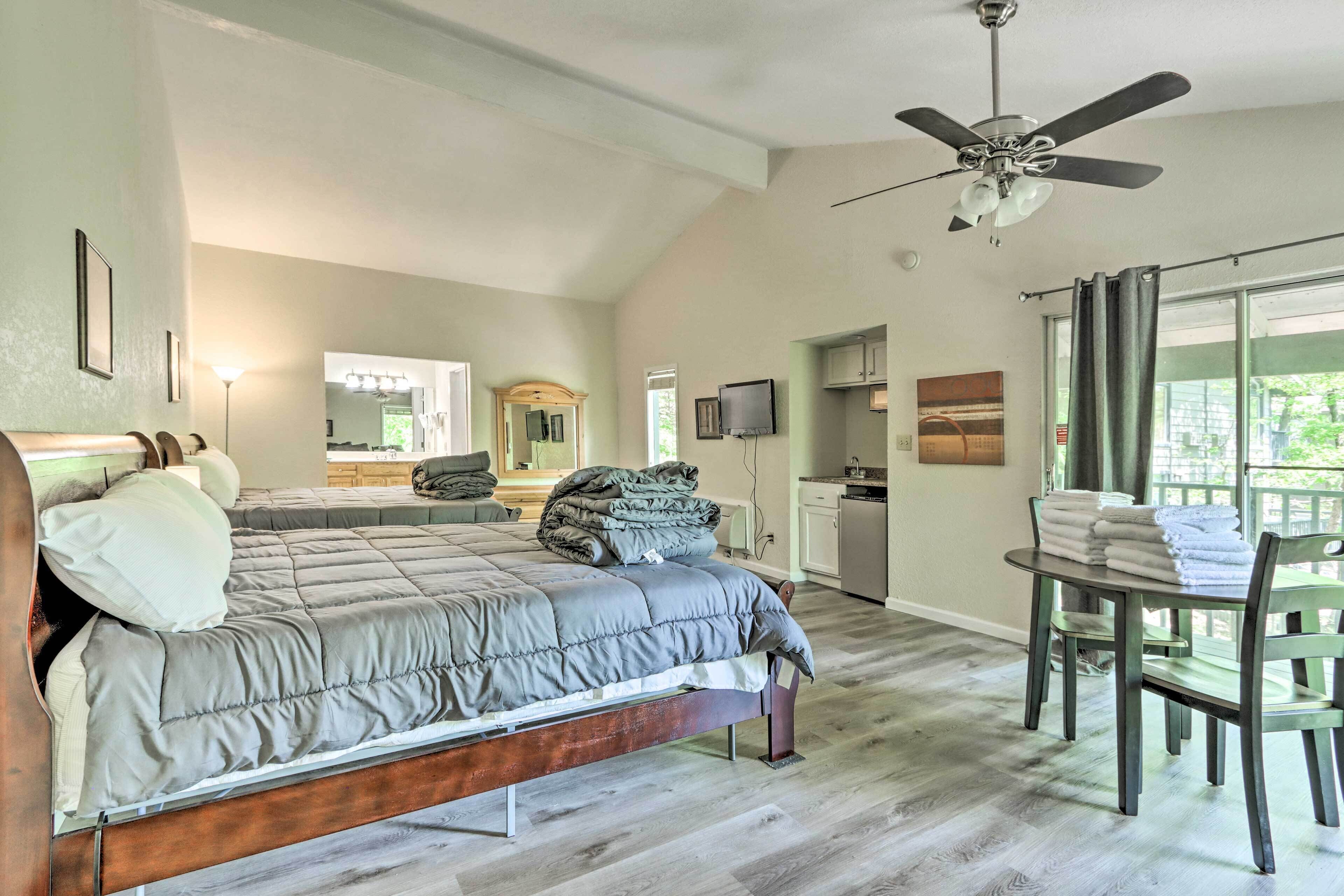 A bedroom with a bed, a ceiling fan, a dining table with chairs, and a view of the outside.
