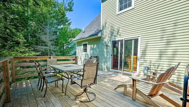 Una terraza de madera con una mesa y sillas, rodeada por una barandilla de madera, y una casa con una ventana y una puerta.