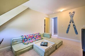 Ein Wohnzimmer mit einem grünen Sofa, einer bunten Decke und einem Couchtisch mit einem Buch. An der Wand hängen Ski und rechts befindet sich ein Fernseher.