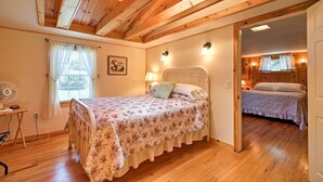 A bedroom with a wooden ceiling, a bed with a floral bedspread, a bedside table with a lamp, and a window with curtains. - 'willow Point' ~ Lake Champlain House w/ 2 Kayaks! (Isle La Motte)