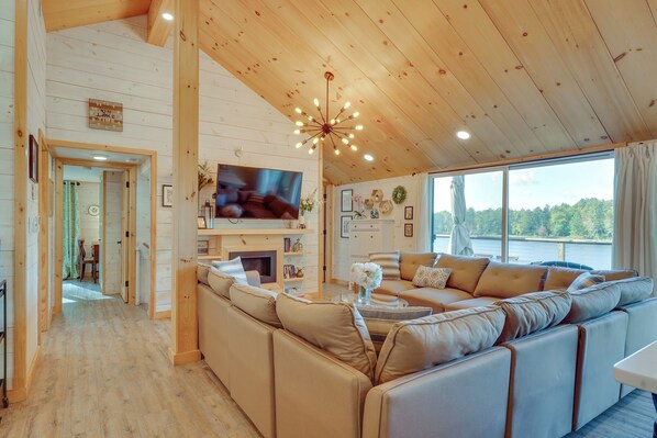 Ein gemütliches Blockhaus-Interieur mit einer großen Holzbalkendecke, einem Fernseher, einem Kamin und Blick auf einen See.