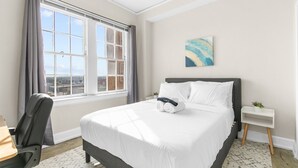 Hotelkamer met een groot raam, een bed met witte lakens, een bureau met stoel, en een klein nachtkastje met een plant.