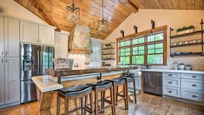 A kitchen with a stone fireplace, stainless steel appliances, wooden bar stools, and a large window with a view of greenery. - Elegant Mtn Chalet w/ Deck: 1 Mi to Mt Snow! (West Dover)