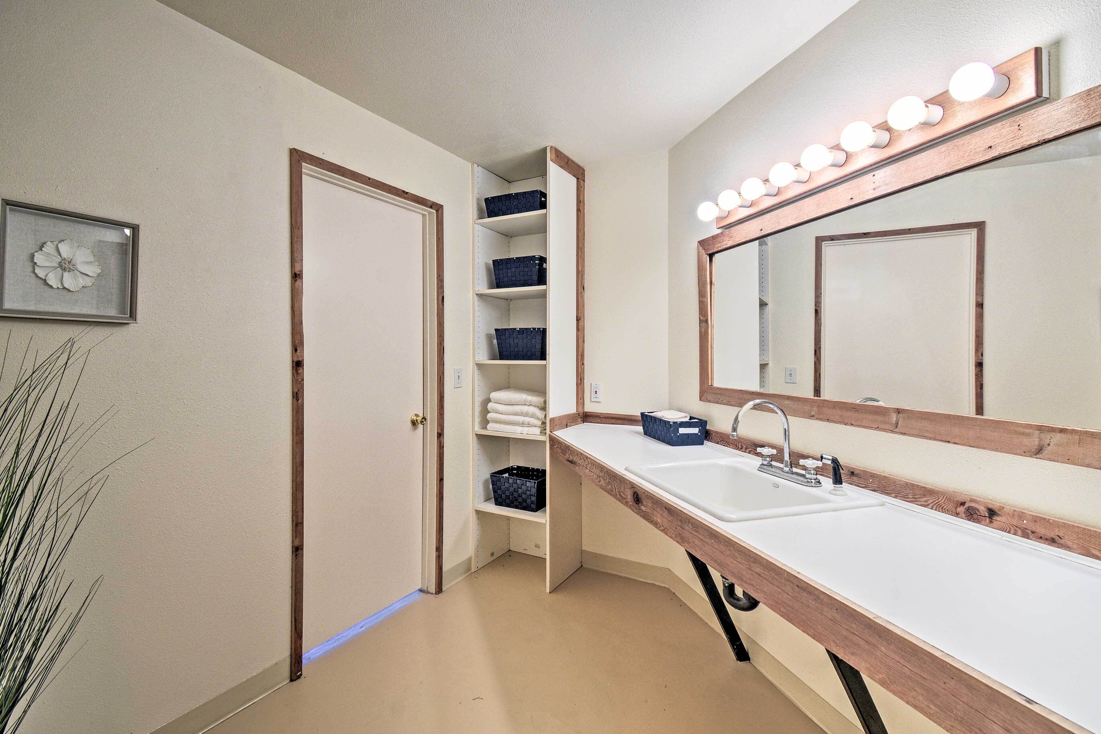 A bathroom with a wooden vanity, a large mirror with lights, and shelves with towels and baskets.