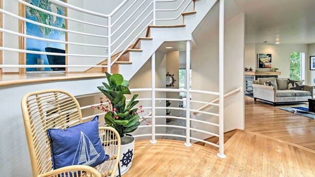 A modern interior with a wooden staircase, a wicker chair with a sailboat pillow, and a potted plant. - Unique Port Ludlow Home w/ 3 Decks & Hot Tub! (Port Ludlow)
