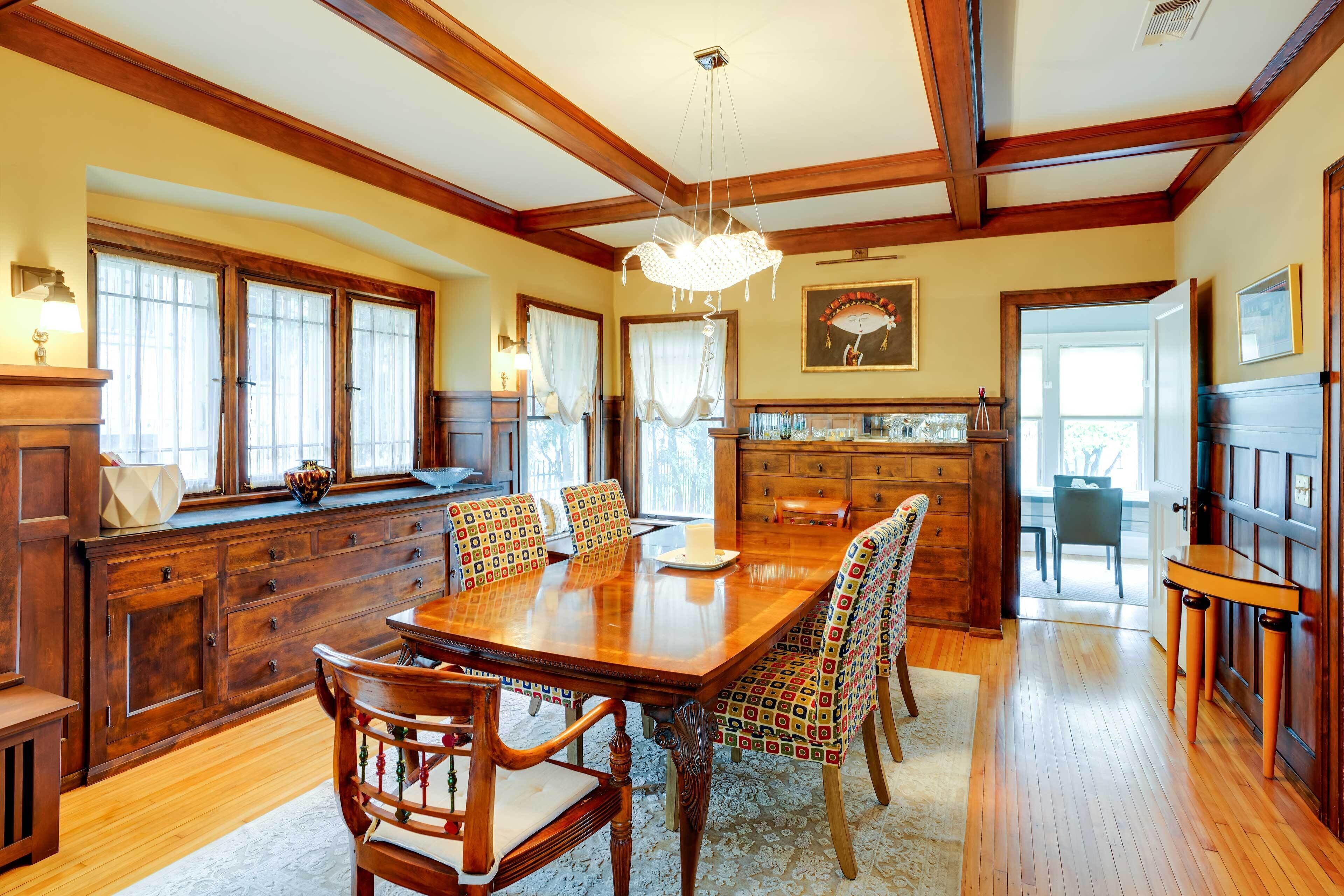 A spacious dining area with a wooden table and chairs, a chandelier, and a view of the kitchen through large windows.