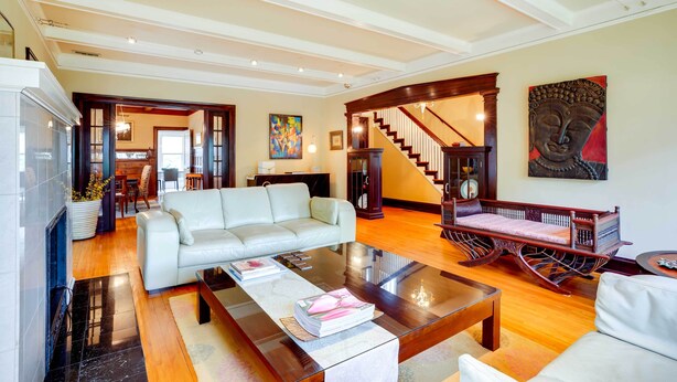 A spacious living room with a white sofa, a glass coffee table, and a wooden chair. A staircase with a wooden railing leads to an upper level.