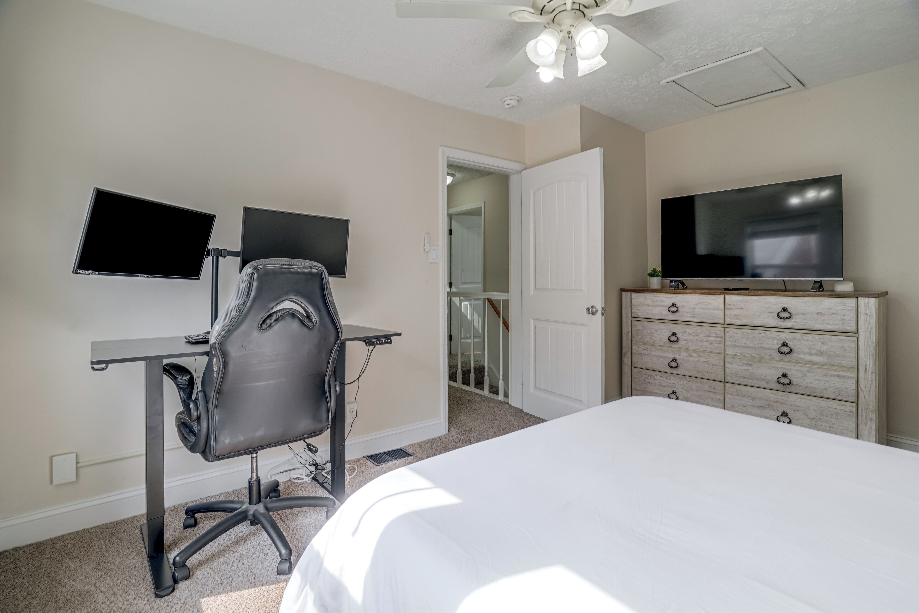 A hotel room with a bed, a desk with two monitors, a chair, a television, and a dresser.