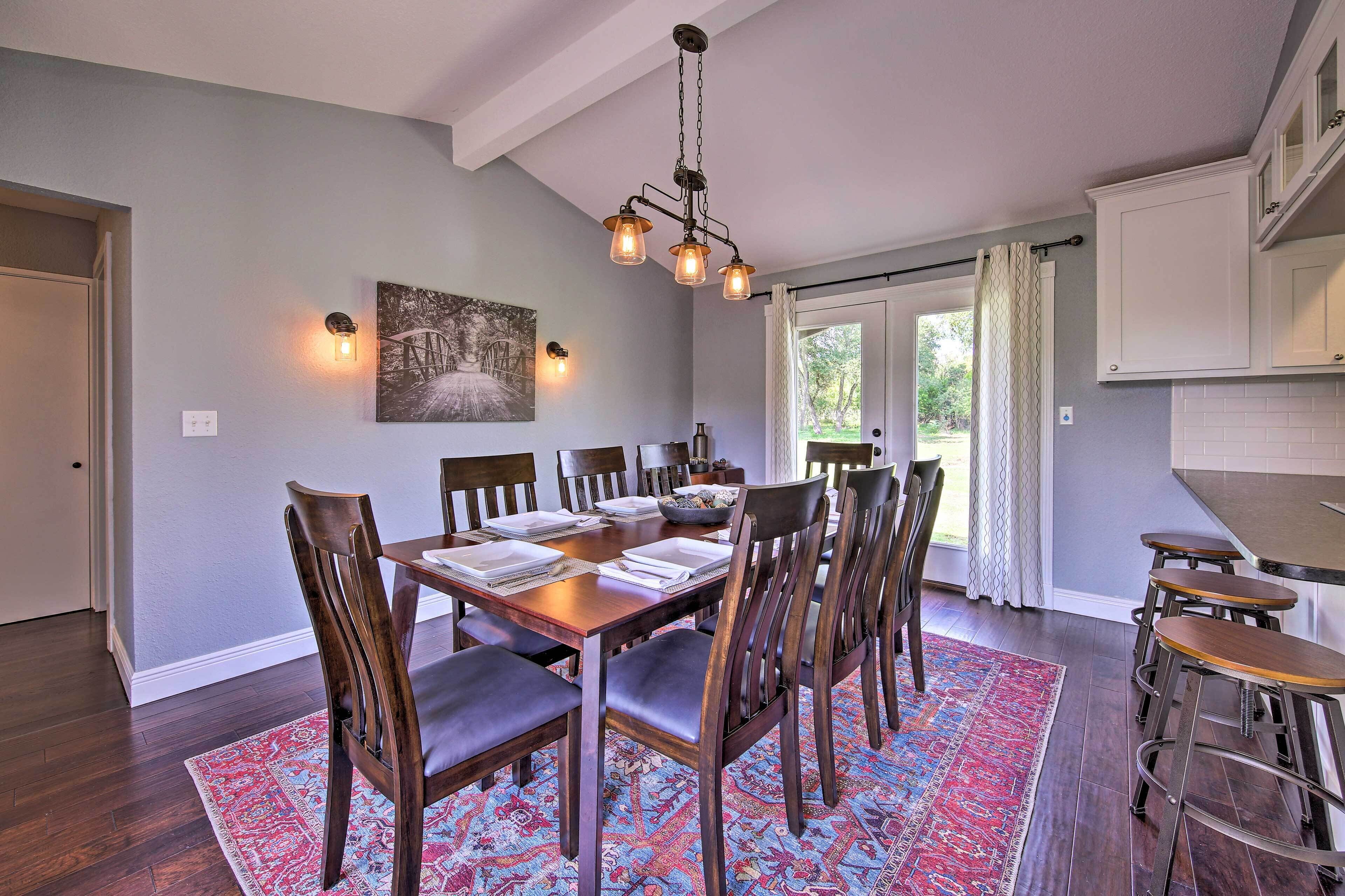 A dining area with a wooden table set for six, surrounded by chairs, a patterned rug, and a hanging light fixture.