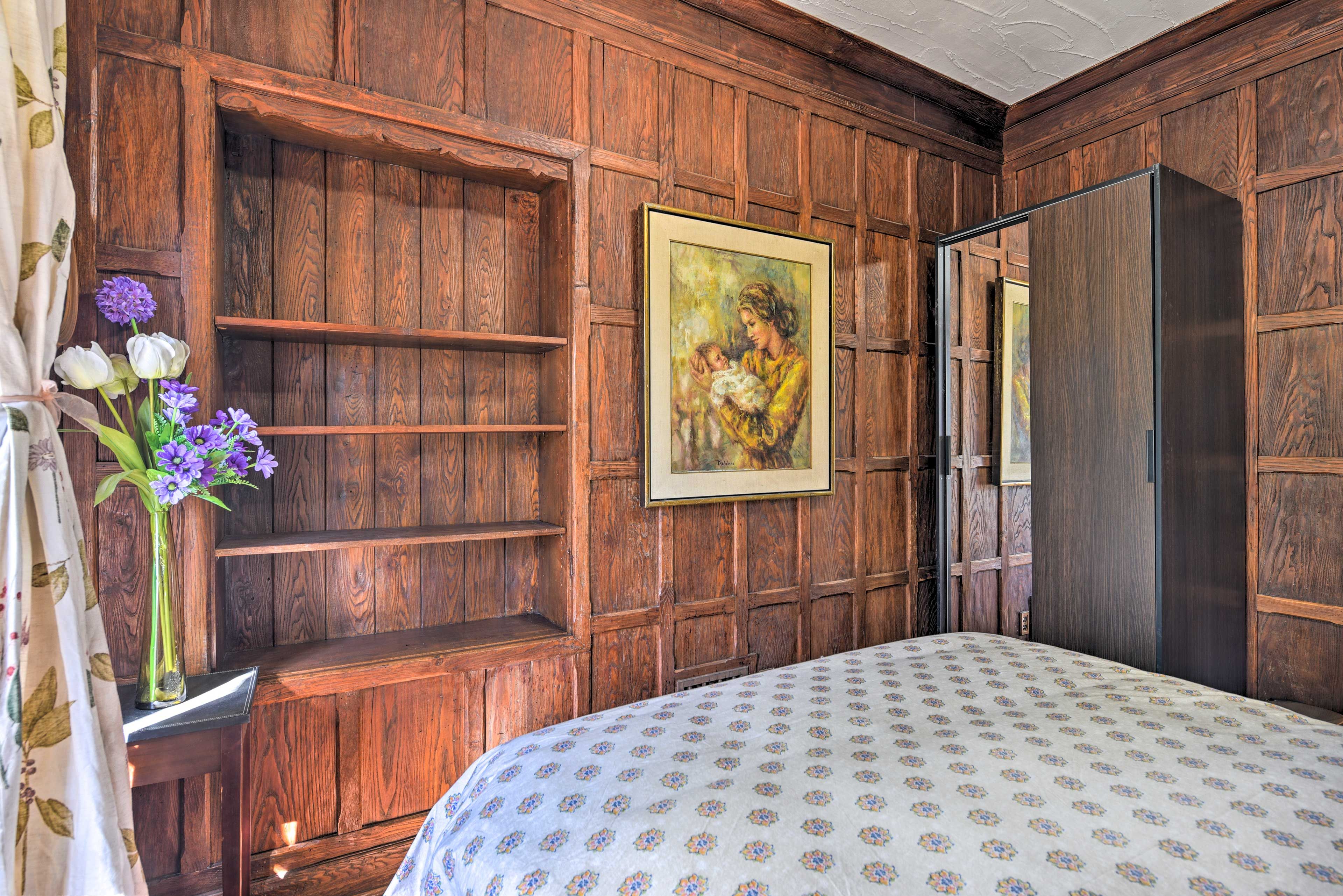 Une chambre avec des lambris en bois, un lit recouvert d’une couvre-lit à motifs, un tableau encadré et un vase rempli de fleurs.