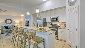 Una cocina moderna con armarios blancos, una isla central con taburetes de bar, electrodomésticos de acero inoxidable, iluminación colgante y un letrero de neón.
