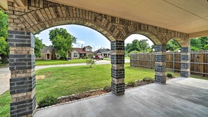 Un porche cubierto con arcos de piedra y piso de concreto, con vista a una zona residencial con casas y una cerca de madera.