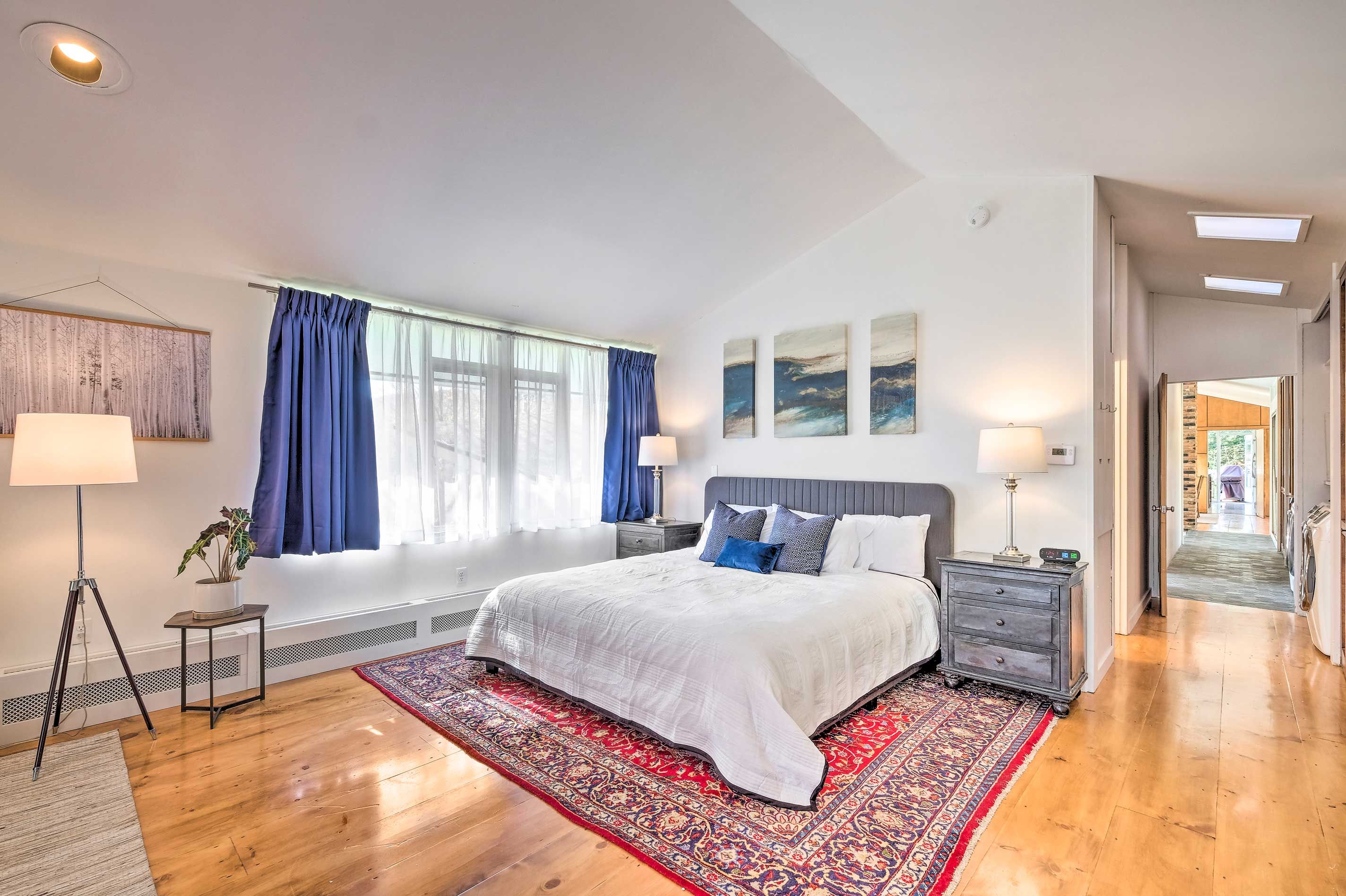 A bedroom with a large bed, two bedside tables, a floor lamp, a standing lamp, a window with curtains, and a view of a garden.