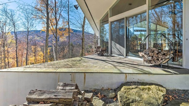 Una casa moderna con un amplio ventanal, una chimenea de piedra y vistas a una cadena montañosa boscosa.