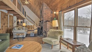 A cozy living room with a fireplace, a high wooden ceiling, and a large window overlooking a snowy landscape.