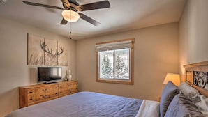 Ein Schlafzimmer mit Bett, Deckenventilator, Fernseher auf einer Holzkommode, Fenster mit Blick auf Bäume und einer Lampe auf dem Nachttisch.