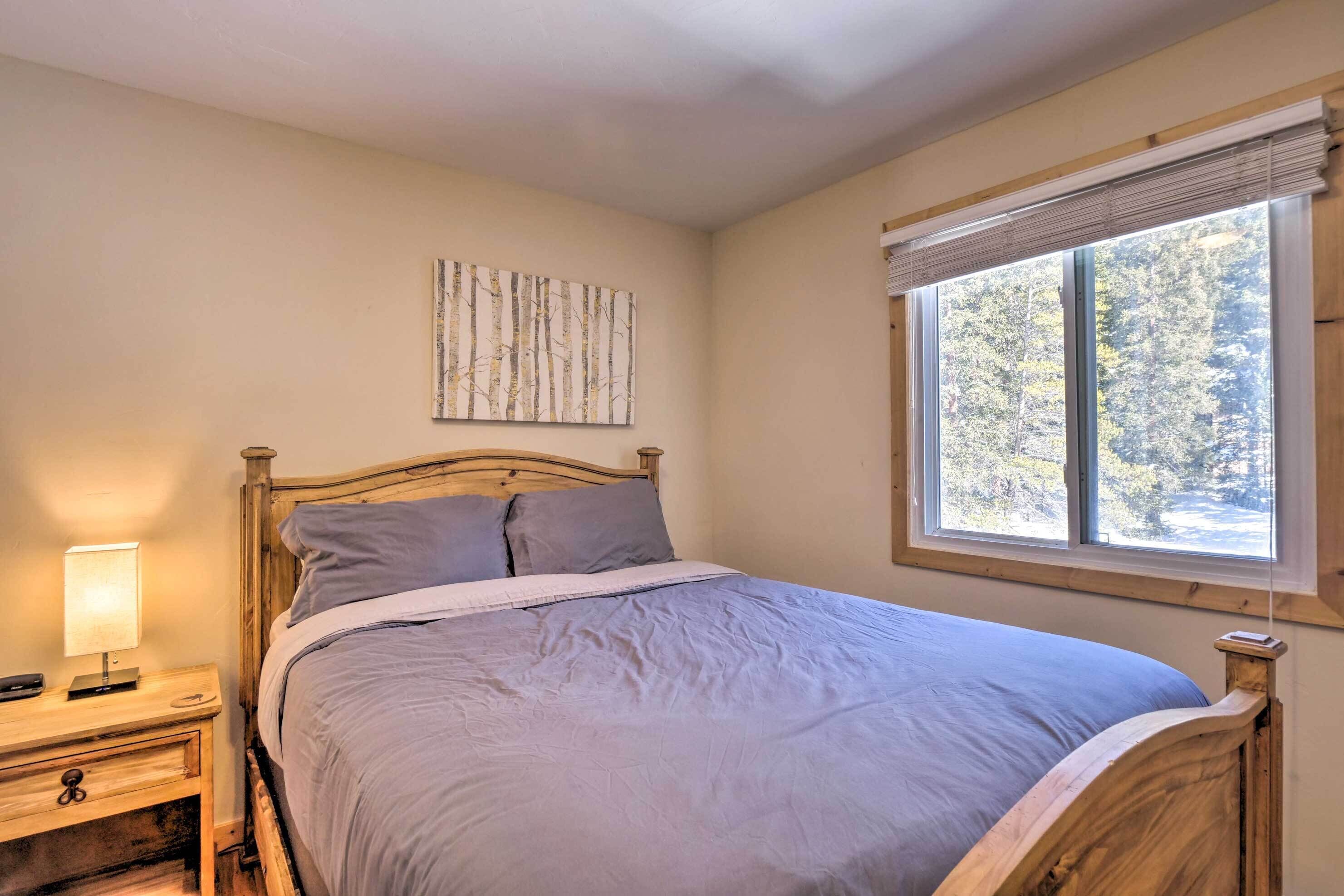 A bedroom with a wooden bed, a bedside table with a lamp, a window with blinds, and a wall-mounted artwork.