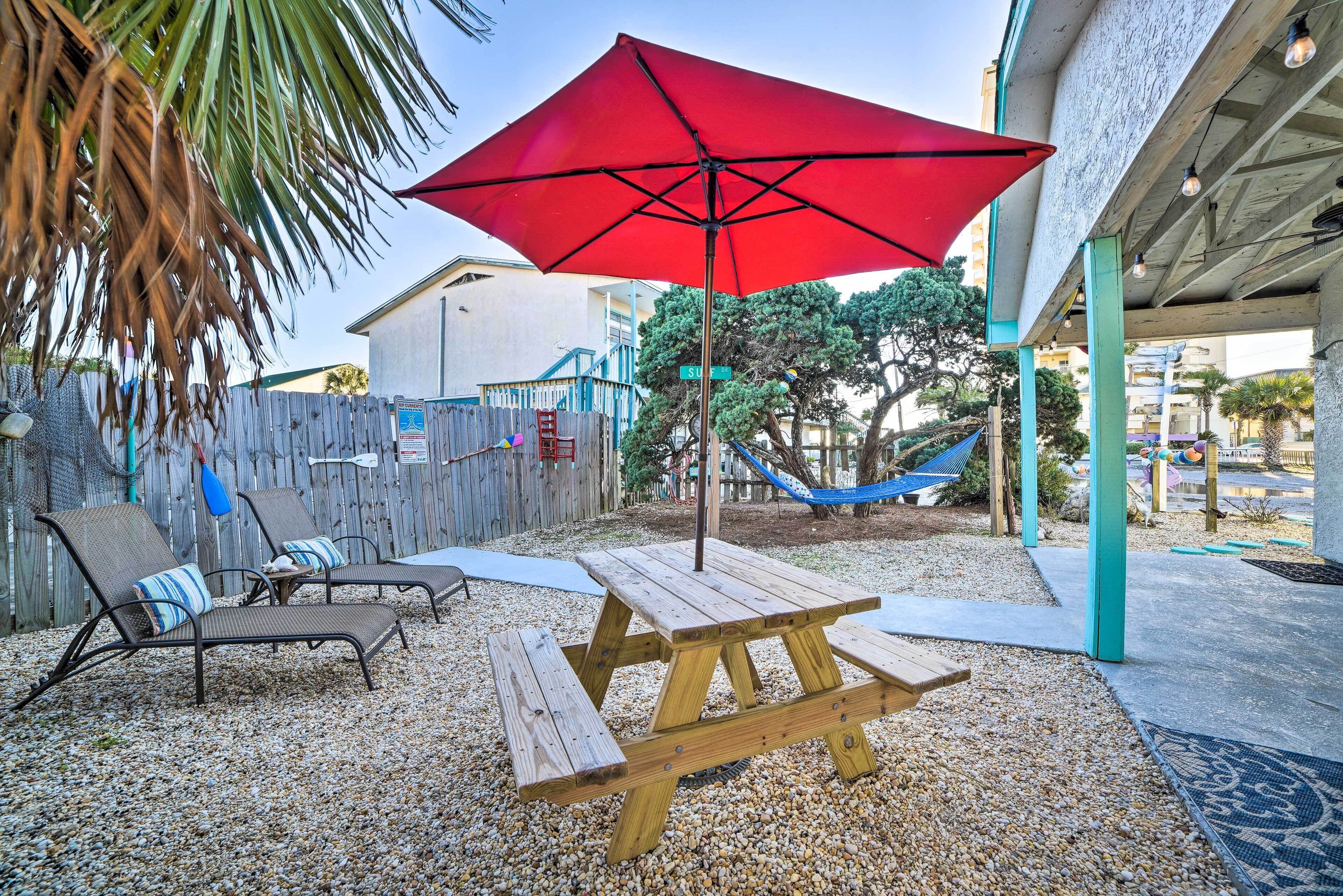 A wooden picnic table with benches and a red umbrella in a backyard with a wooden fence, a hammock, and a view of a building in the background.