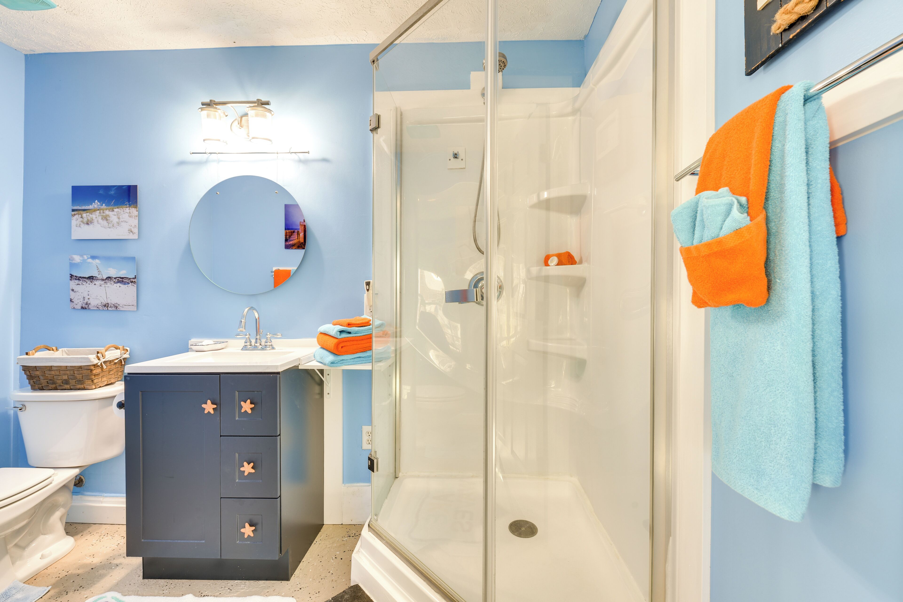 Une salle de bain avec une douche à parois de verre, des toilettes blanches, un meuble-lavabo de couleur foncée doté de poignées en forme d’étoile, et un miroir rond au-dessus du lavabo.