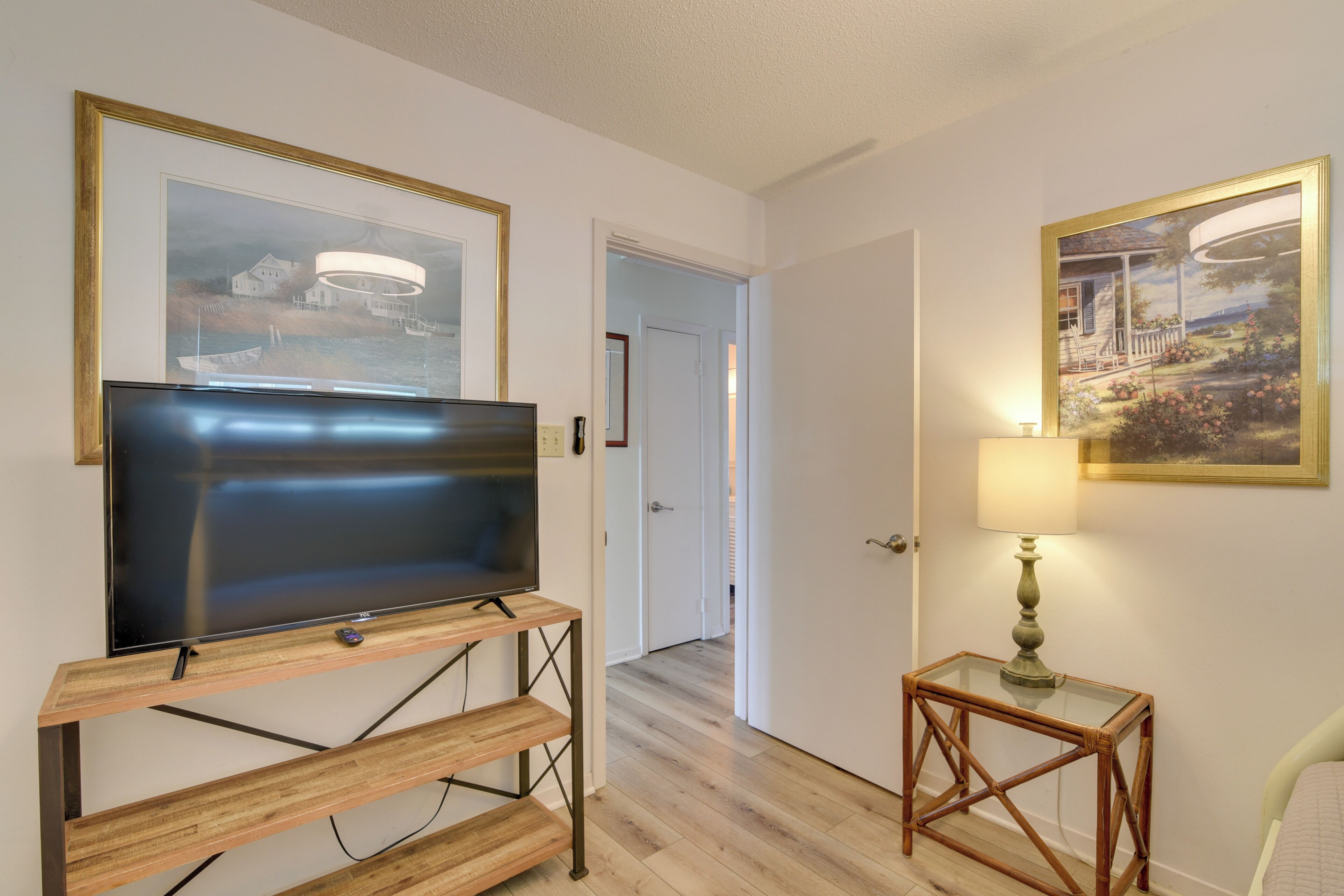 A room with a flat-screen TV on a wooden stand, a small table with a lamp, and two framed pictures on the walls.