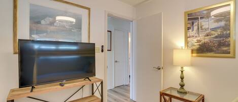 A room with a flat-screen TV on a wooden stand, a small table with a lamp, and two framed pictures on the walls.