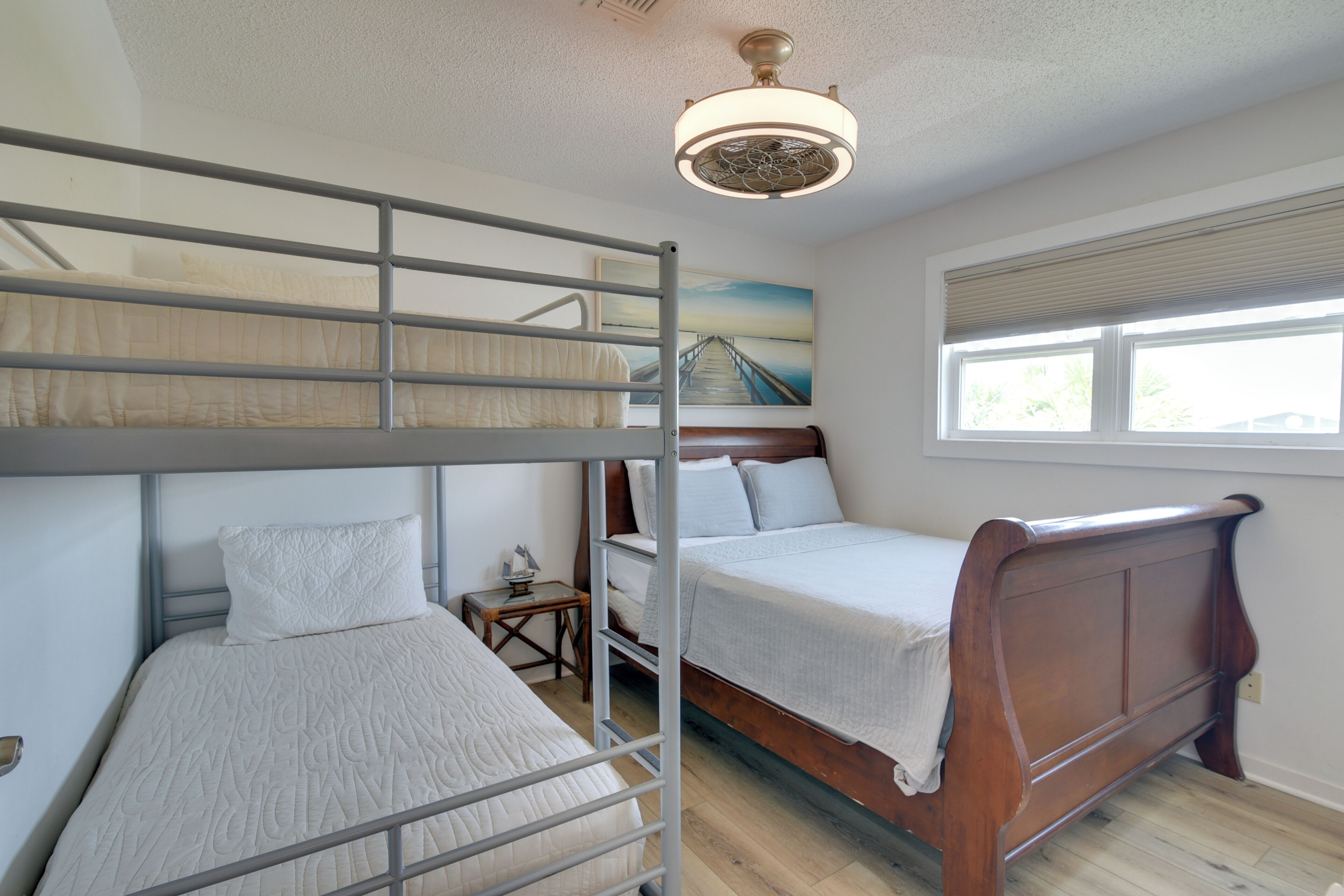 A room with bunk beds, a small table, a nightstand, a ceiling fan, and a window with blinds.