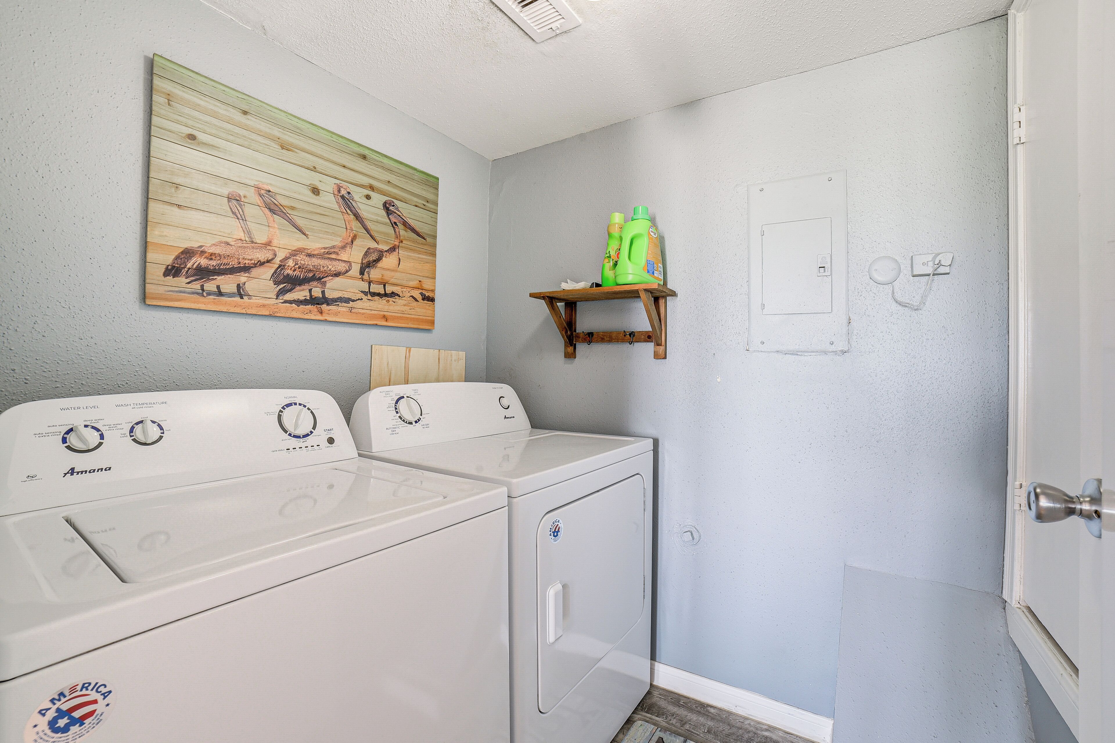 A laundry area with two white Amana washers and dryers, a wooden shelf with green cleaning products, and a wall-mounted electrical box.