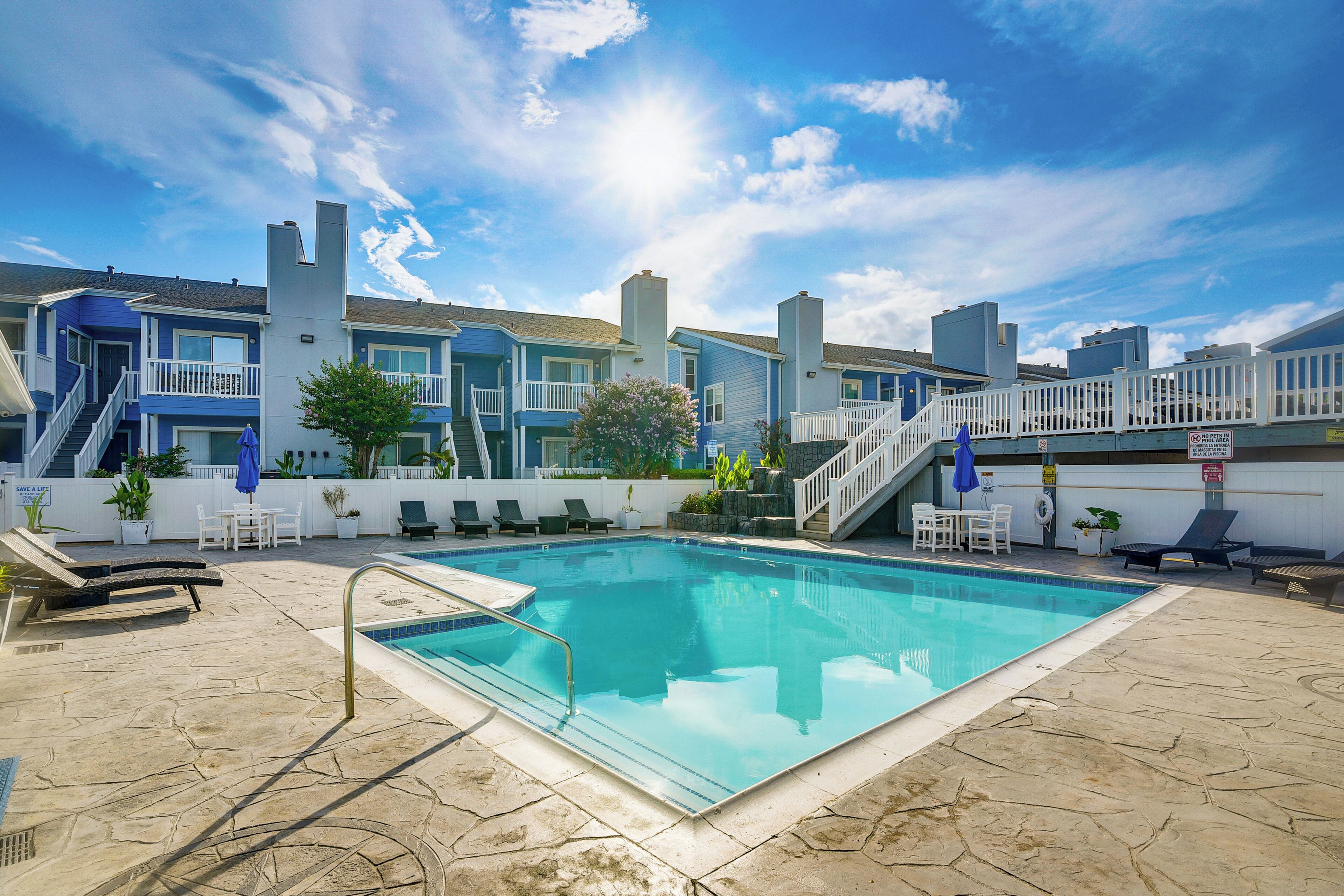 A swimming pool with a stone deck, lounge chairs, and a staircase leading to a building with blue and white exterior walls.