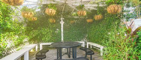 An outdoor patio with a round table and benches, surrounded by hanging planters and a white lattice structure.
