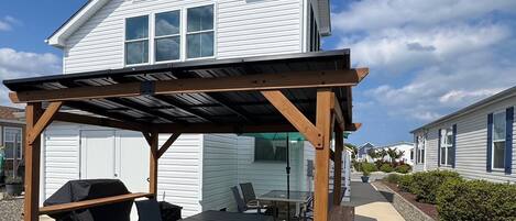 A covered patio with outdoor furniture, a grill, and a house in the background.