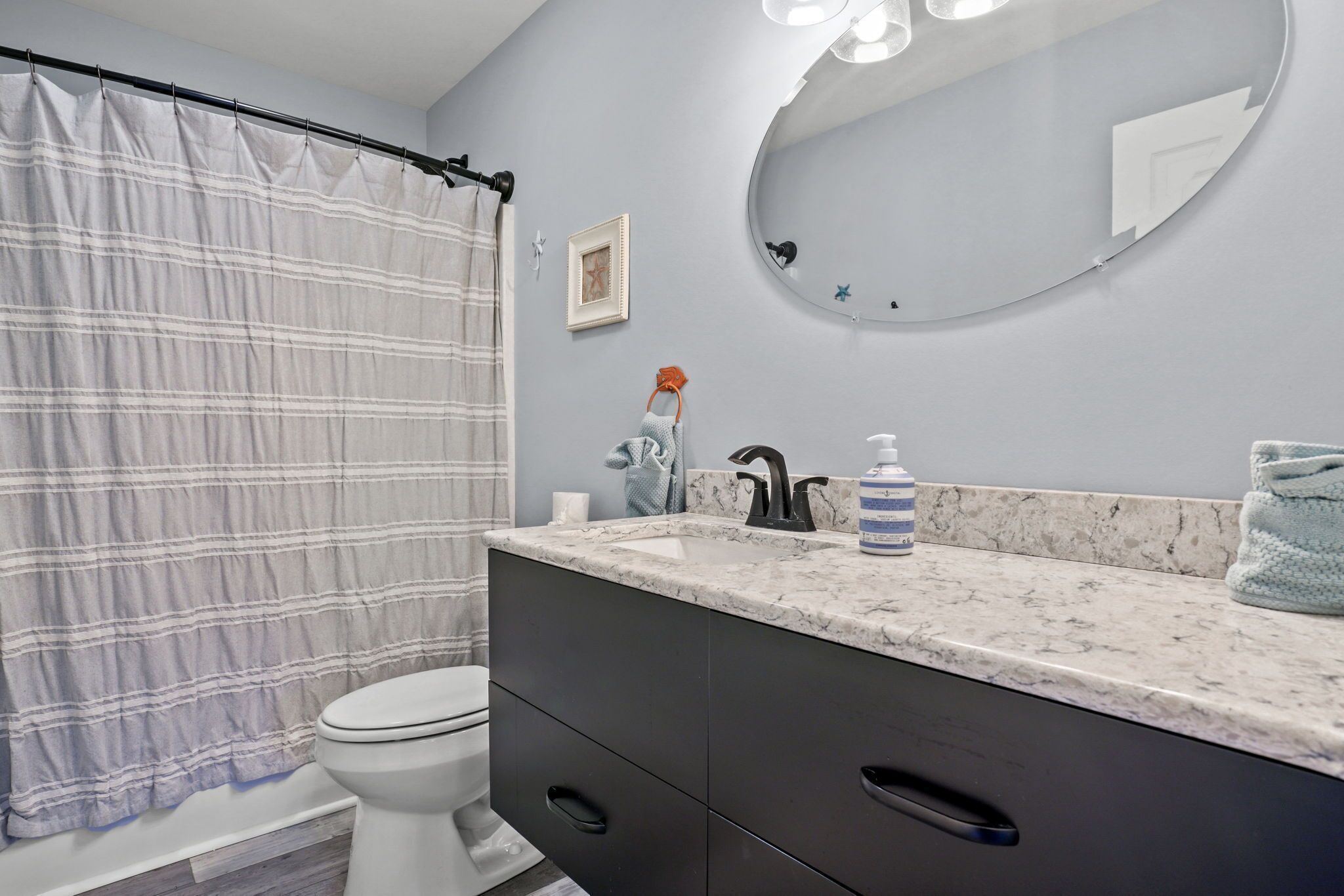 A bathroom with a striped shower curtain, a white toilet, a sink with a marble countertop, and a round mirror.