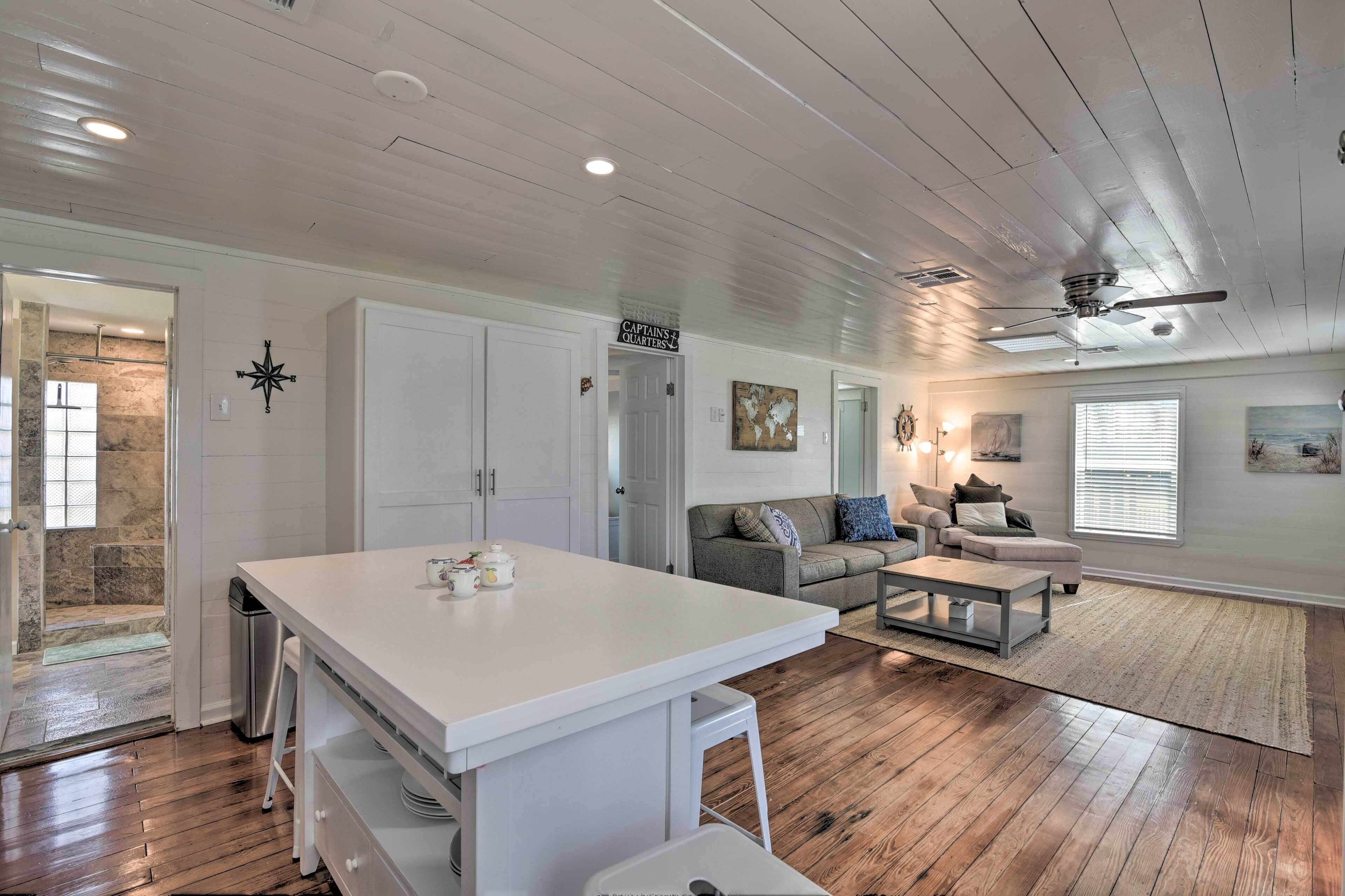 A spacious living area with a kitchen island, wooden flooring, a ceiling fan, and a view into a stone-walled bathroom.