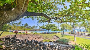 Un jardín tropical con un gran árbol, un área pavimentada con rocas y vista al mar y a la playa en el fondo.