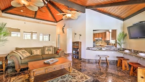 A spacious living area with a wooden ceiling, a sofa set, a coffee table, and a kitchen area with a TV and bar stools.