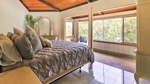A bedroom with a large bed, a ceiling fan, a window with a view of palm trees, and a wooden ceiling.