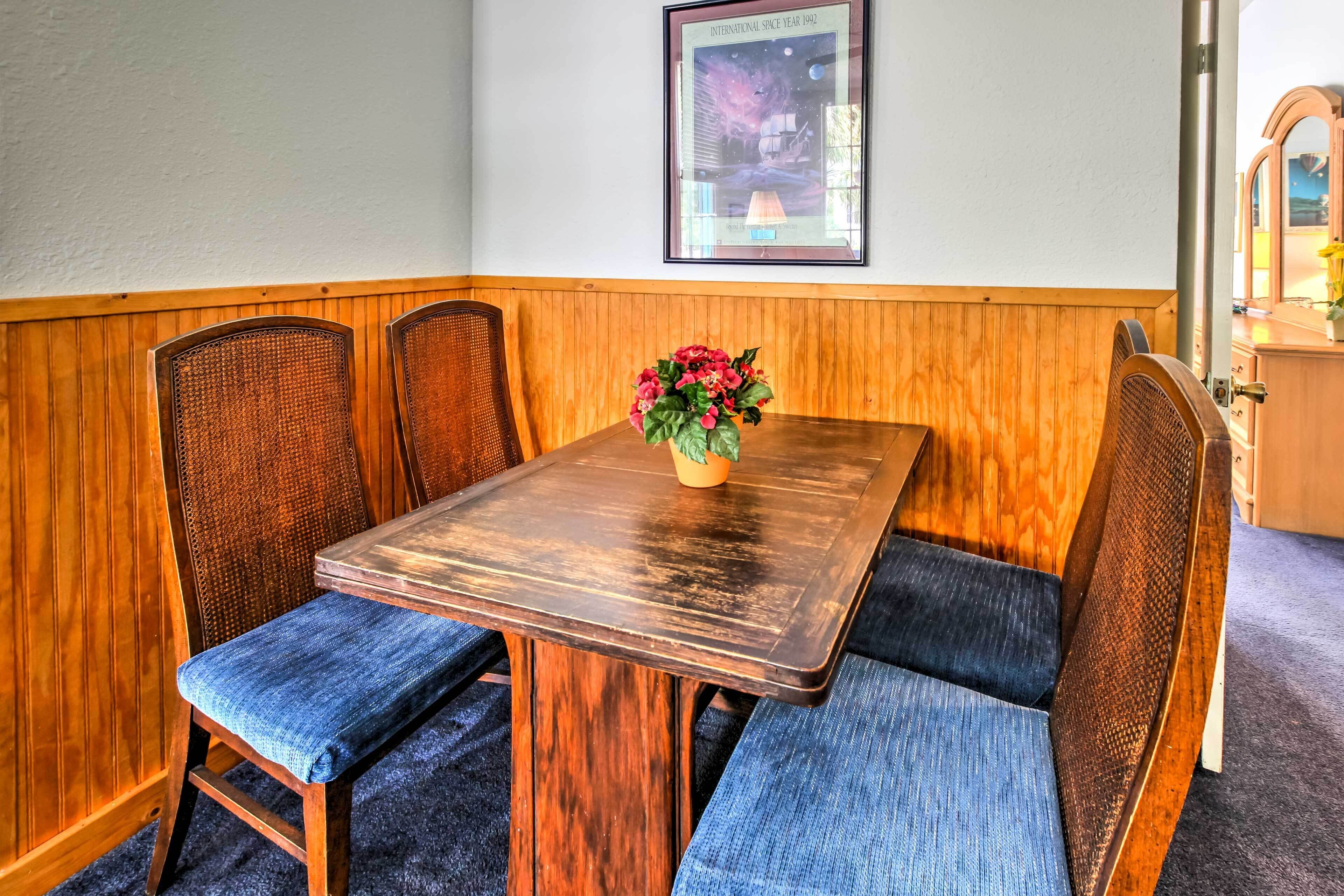 A corner seating area with a wooden table and blue cushioned chairs, a framed picture on the wall, and a wooden cabinet with a mirror.